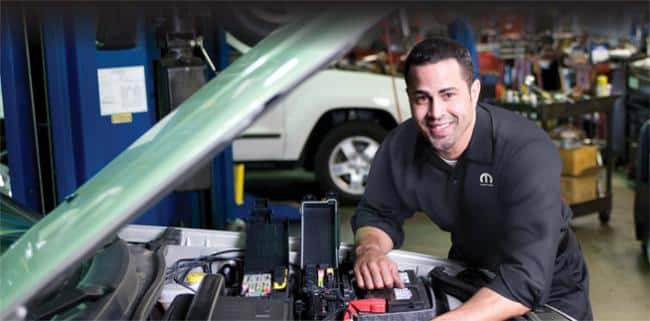 Mechanic smiling while working on a car engine in a busy workshop.